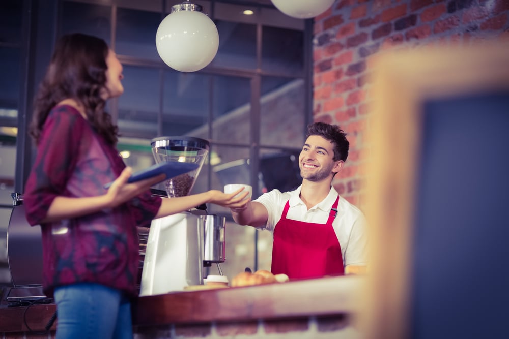 Barista at cafe hands customer a drink as she holds a tablet in her hand.