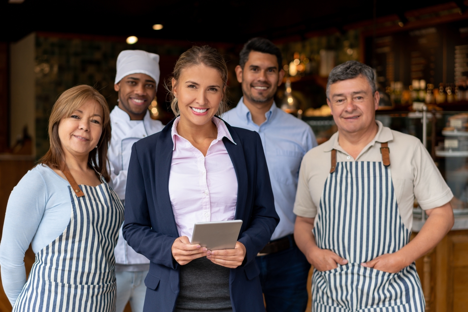 Restaurant manager holding a tablet standing with a smiling team of chefs and staff discussing the daily log and shift log.