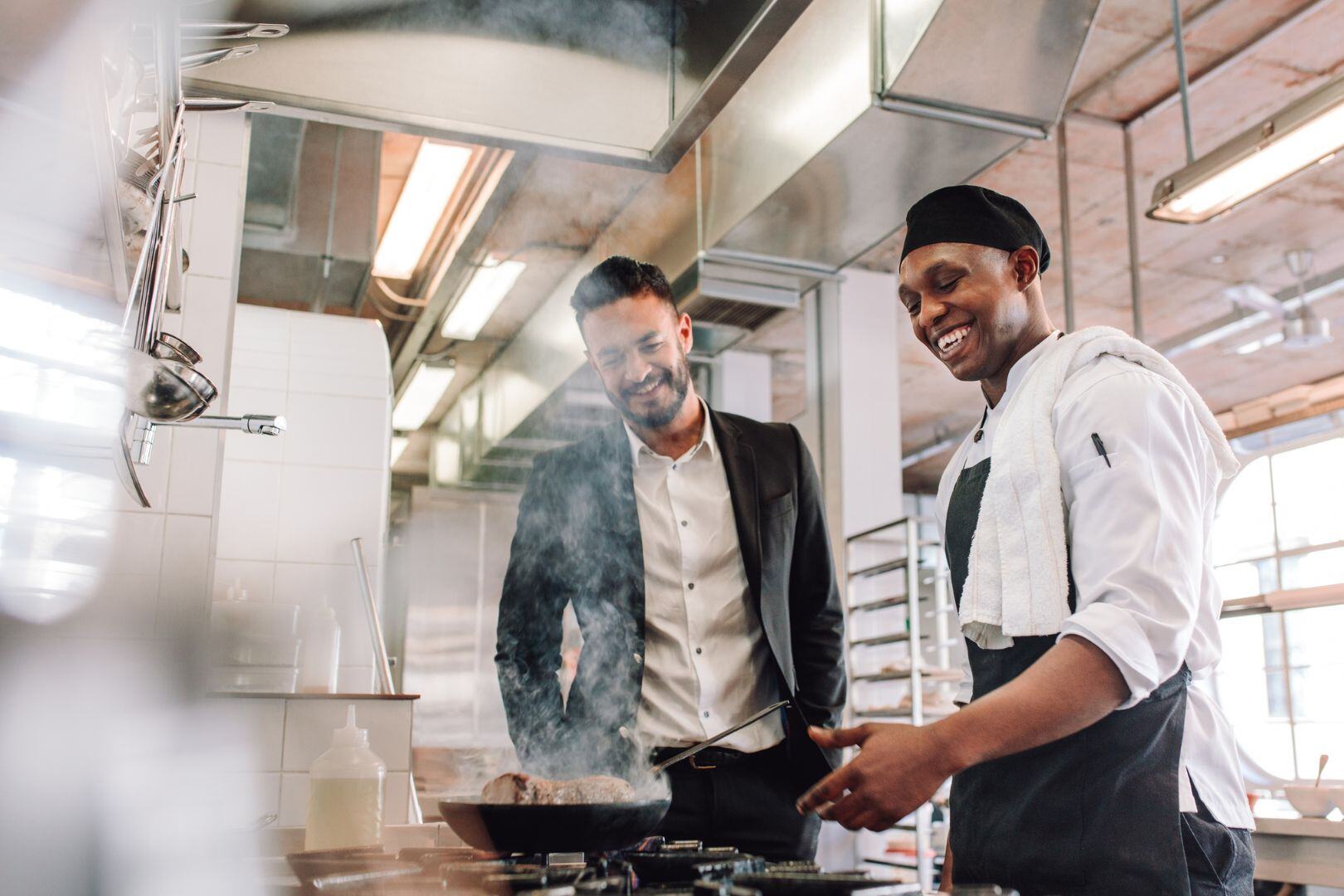 Restaurant manager talking with a smiling chef while cooking in a busy commercial kitchen.